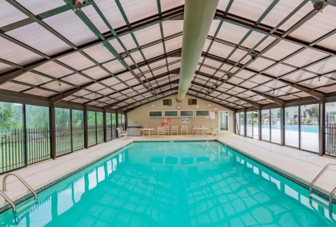 An indoor pool area features a spacious swimming pool with clear turquoise water. Sunlight filters through expansive glass panels, providing a bright ambiance. Surrounding the pool are rows of chairs, while transparent walls offer views of the outdoor area, enhancing the relaxing atmosphere.