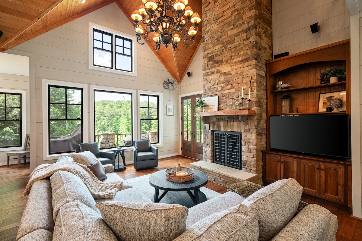 A spacious living area showcases a stone fireplace as a focal point, surrounded by comfortable seating arrangements. Large windows allow natural light to fill the space, offering views of the surrounding greenery. A chandelier hangs above, enhancing the inviting atmosphere of the room.