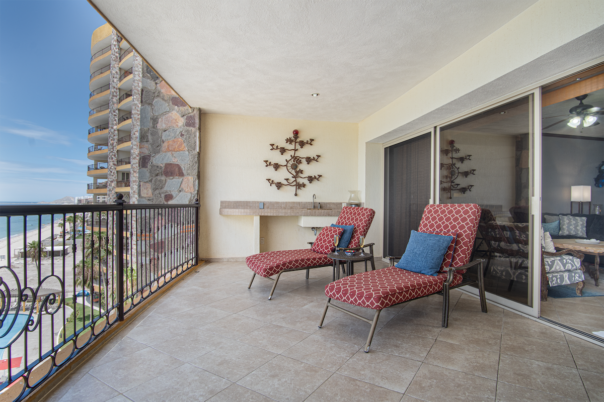A spacious balcony area features two lounge chairs with patterned upholstery, positioned to overlook the ocean. A stone wall provides a backdrop, and a small sink is integrated into the design. Large sliding glass doors lead to the indoor living space, inviting natural light.