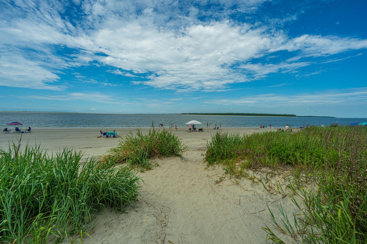 The image captures a serene beach scene with gentle waves lapping at the shore under a bright blue sky. A sandy path lined with lush green grass leads to the beach, where sunbathers and umbrellas dot the landscape, inviting relaxation and enjoyment by the water.
