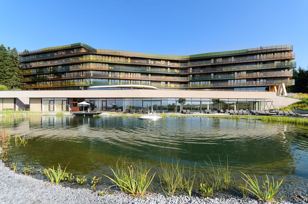 The contemporary hotel structure is set against a clear blue sky, featuring layered balconies and expansive glass windows. A serene pond with grasses and small plants surrounds the building, creating a tranquil atmosphere. Lounge chairs line the space for relaxation.
