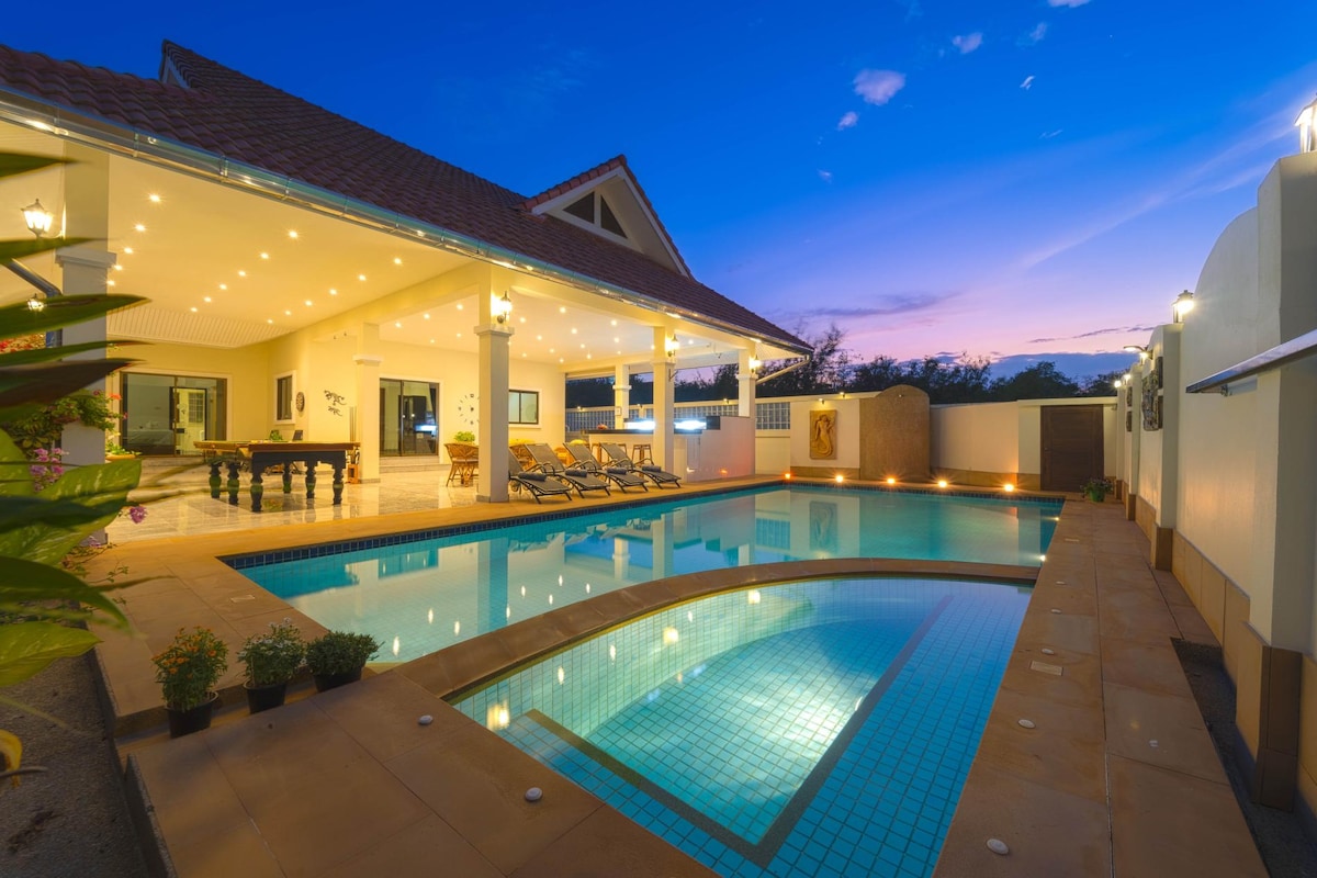 An expansive outdoor area features a large swimming pool with a tiled surface. Comfortable sun loungers line the pool deck under soft evening lighting. The spacious villa is shown with multiple indoor and outdoor seating areas, creating a welcoming environment for relaxation.