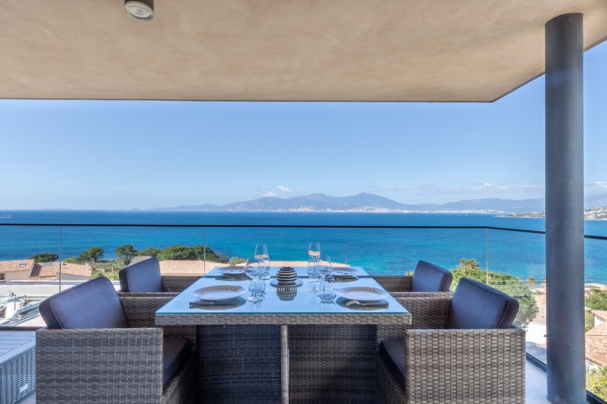 A dining area is presented on a spacious terrace, featuring a glass-top table surrounded by woven seating. The sea and distant mountains are visible in the background, enhancing the outdoor setting. Clear skies and calm waters contribute to a serene atmosphere.