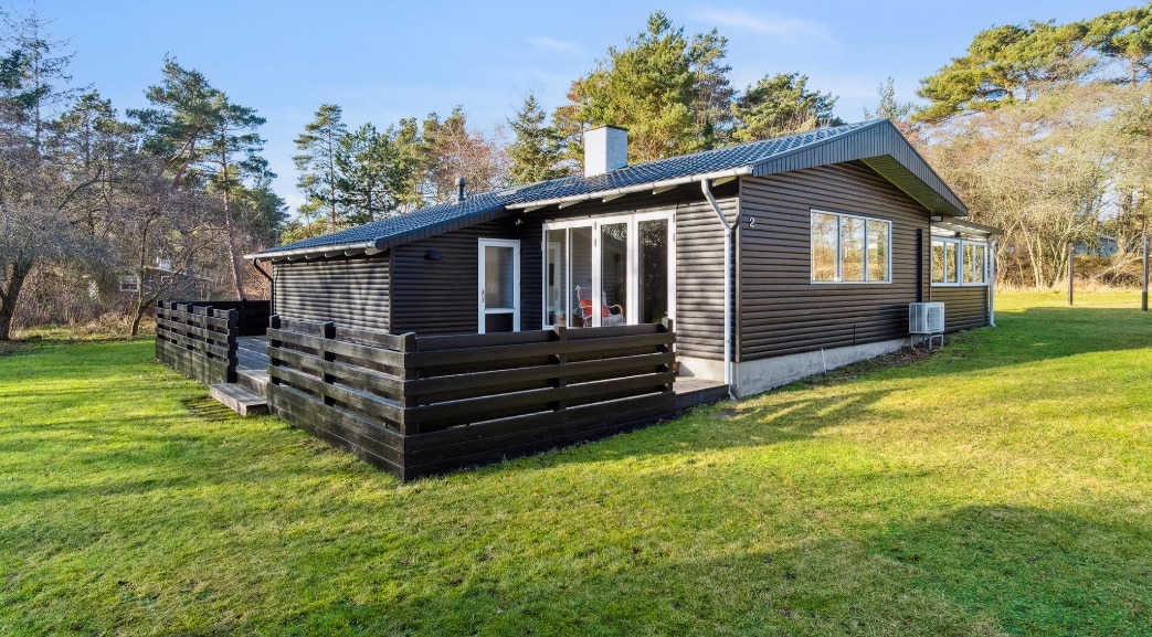 The summer cottage features a wooden exterior with large windows, allowing natural light to fill the interior. A spacious deck surrounds the front, framed by low wooden railings. The extensive lawn is bordered by trees, enhancing the sense of privacy and space.