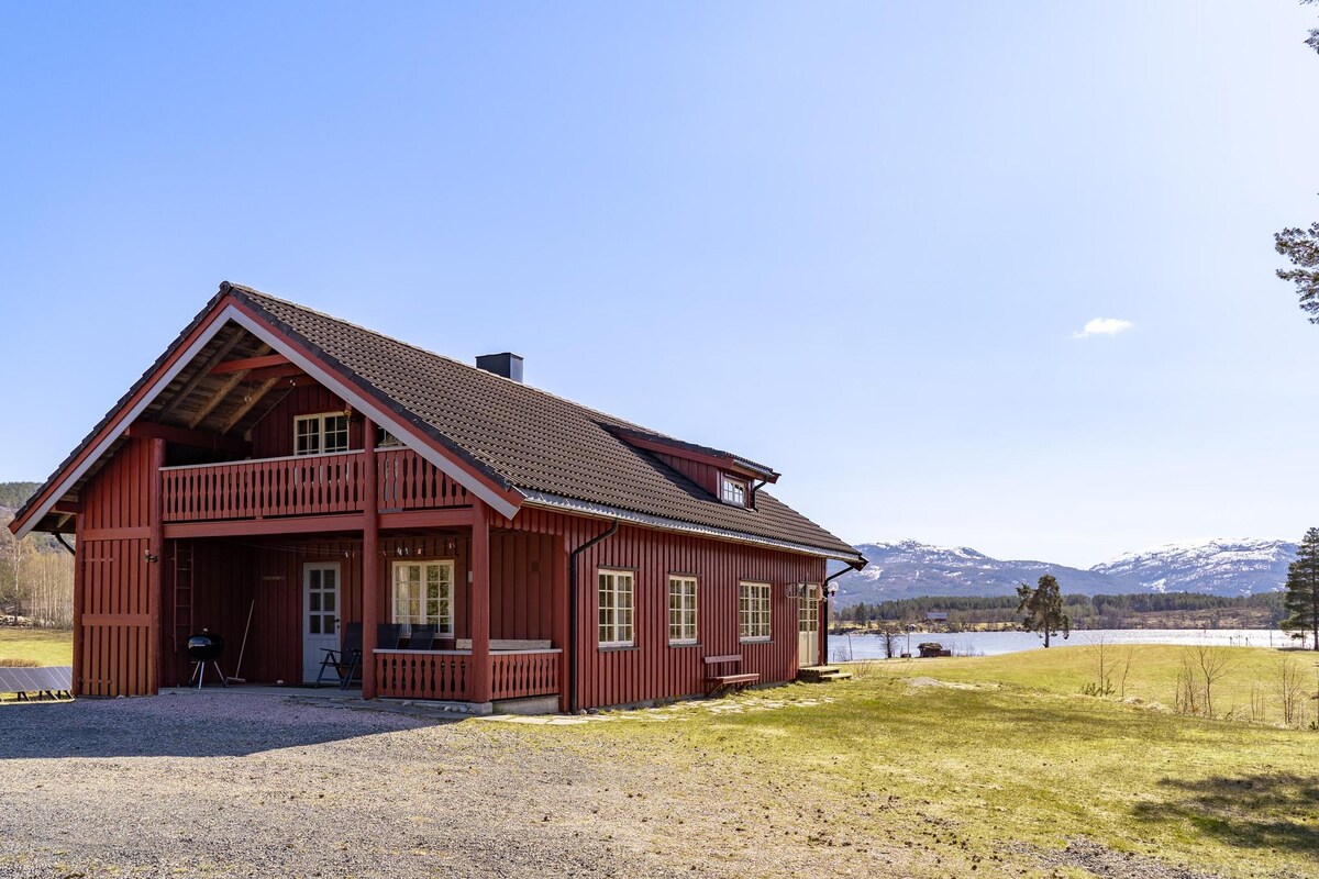 The charming wooden house is set against a backdrop of rolling hills and a clear blue sky. A large grassy area surrounds the property, enhancing the outdoor space. Features include a balcony and outdoor seating, providing an inviting view of the nearby lake.