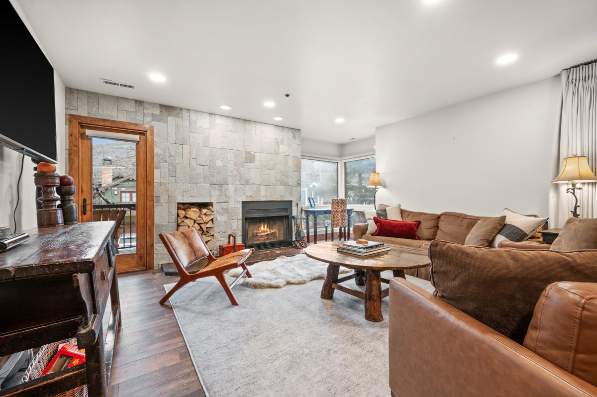 A cozy living room features a stone fireplace, surrounded by comfortable seating and a large coffee table. Natural light enters through multiple windows, illuminating the warm tones of the furnishings and enhancing the inviting ambiance.