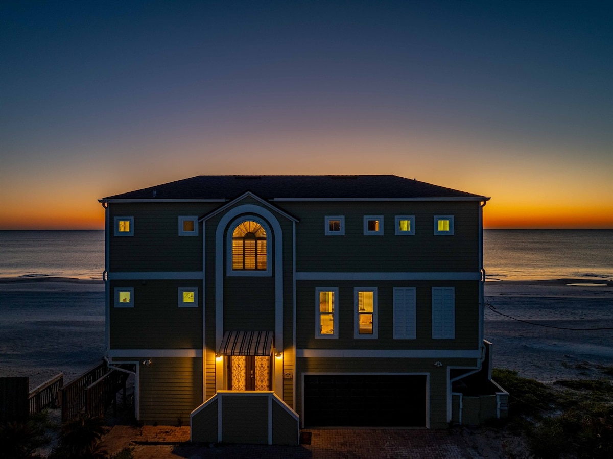 The three-story coastal retreat is illuminated at dusk, showcasing its welcoming entrance and multiple windows. The vibrant colors of the sunset reflect on the ocean, creating a serene backdrop. The property is positioned just steps from the beach, enhancing its beachside appeal.