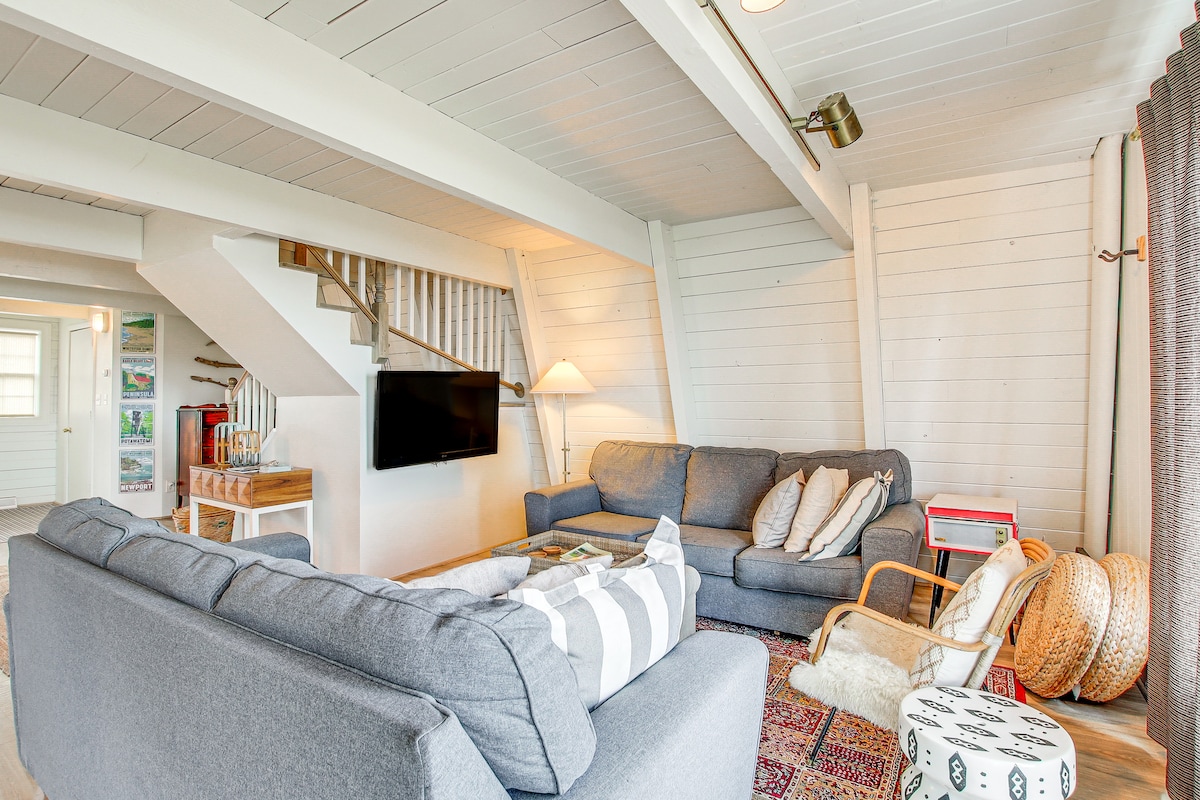 An inviting living area features a two-tone sofa and a cozy armchair, arranged around a central coffee table. A Smart TV is mounted on a white wooden wall. Light streams in from a nearby window, while a decorative rug adds warmth to the space.