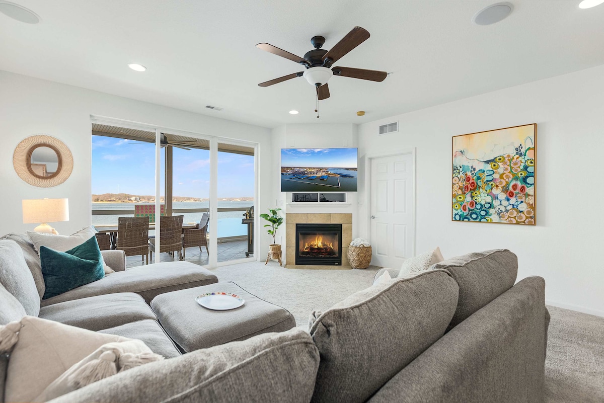 A spacious living room features a gray L-shaped sofa, a mounted smart TV, and a cozy fireplace. Large sliding doors provide access to a balcony, while decorative artwork and natural light enhance the inviting atmosphere.
