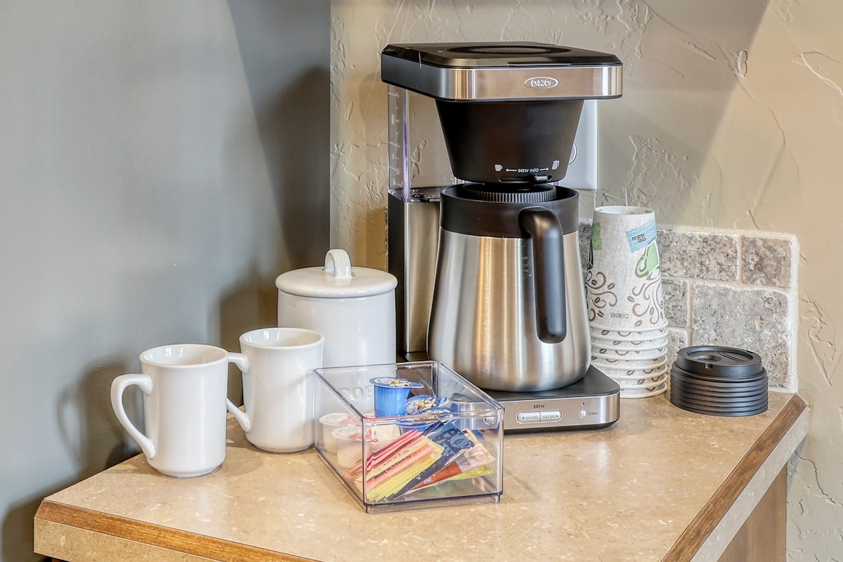 A coffee station is presented on a countertop, featuring a stainless steel coffee maker alongside two white mugs. A white sugar container and a clear box filled with assorted coffee pods and condiments sit nearby, providing convenience for guests.