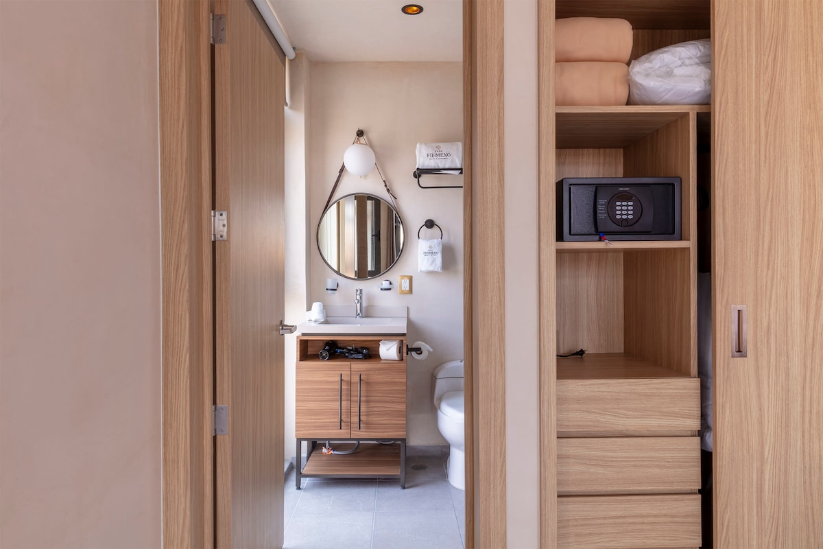 A well-organized bathroom area is visible, featuring a wooden vanity with a round mirror above it. Clean towels and toiletries are neatly arranged. A storage unit beside the vanity includes a safe, while a toilet is positioned in the background.