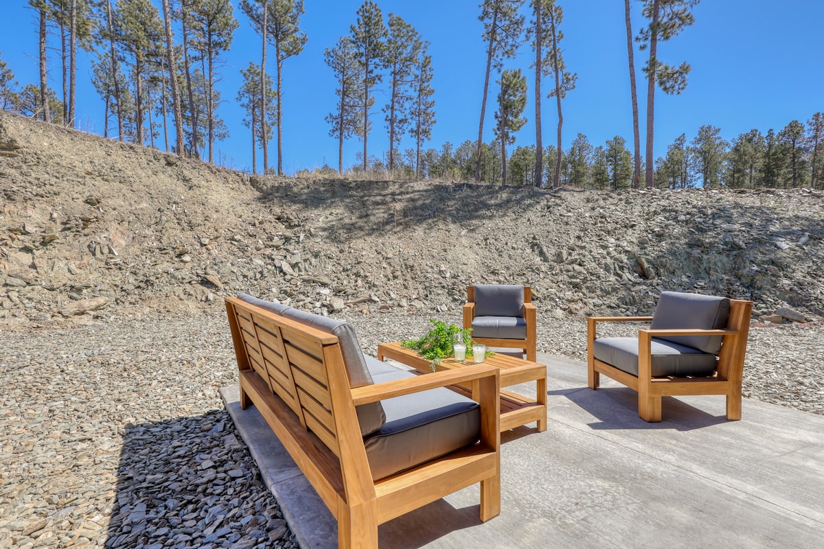 An outdoor seating area features a wooden couch and two armchairs with gray cushions, arranged around a small table. The backdrop consists of rocky terrain and tall pine trees under a clear blue sky.