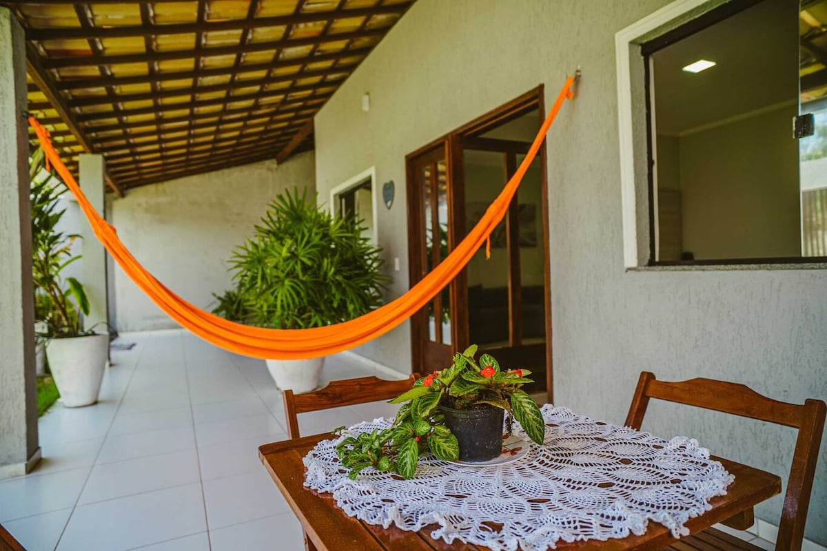 An outdoor terrace features a wooden dining table adorned with a lace tablecloth and a potted plant. An orange hammock is suspended nearby, providing a comfortable spot for relaxation. The covered area is bordered by greenery and large windows, enhancing the inviting atmosphere.