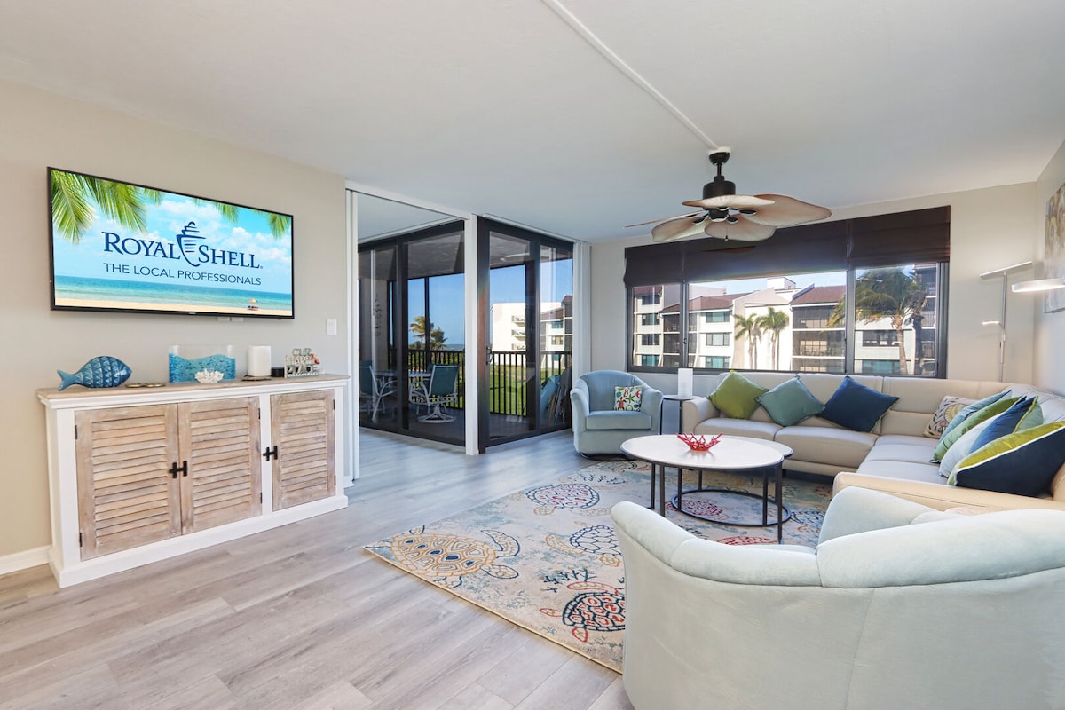 A comfortable living area features a light-colored sofa adorned with colorful throw pillows. A circular table sits in front of the sofa, and a flat-screen TV is mounted on the wall. Large windows provide natural light, showcasing views of the screened lanai.