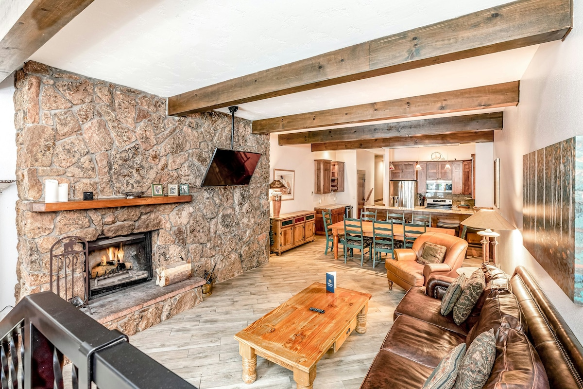 The living room features a stone fireplace with a wooden mantel, paired with a leather sofa and overstuffed chairs. A wooden coffee table sits in front of the seating area. A wall-mounted TV is positioned for easy viewing, while an open kitchen and dining space are visible in the background.