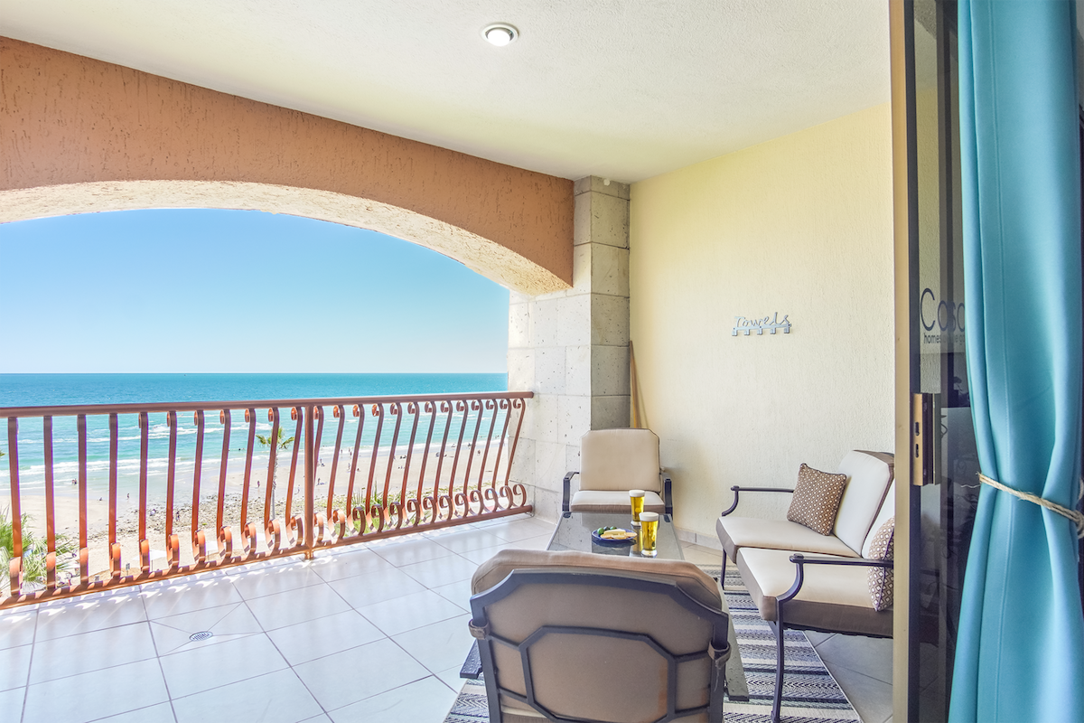 A private patio area features comfortable seating arrangements overlooking the beach. The space is complemented by a refreshing coastal view and a gentle sea breeze, visible through the sliding glass doors that connect to the living room.