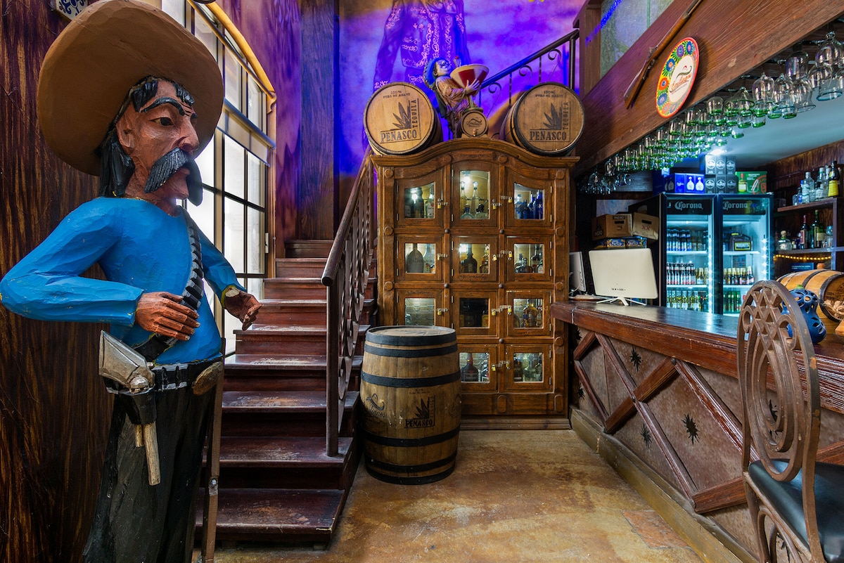 A rustic bar area features wooden shelves adorned with glassware and bottles. The warm tones of the wood contrast with vibrant murals. A life-sized statue of a figure in traditional attire stands near the entrance, adding character to the welcoming space.