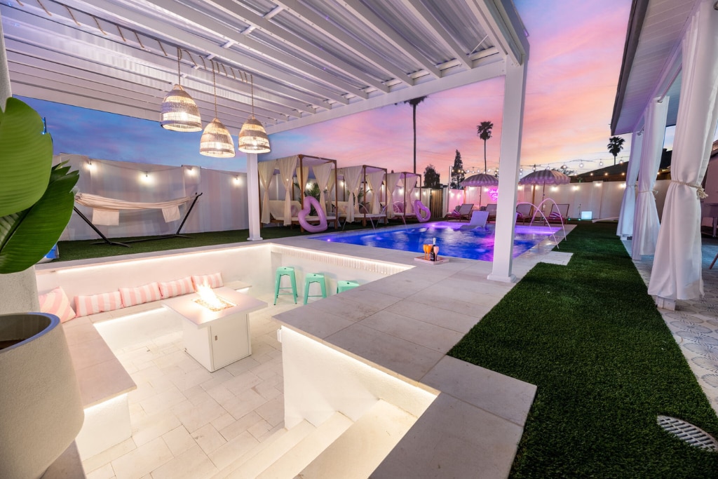 An inviting outdoor space features a heated pool surrounded by lush greenery and vibrant lounge chairs. A sunken seating area includes a table and fire feature, while stylish light fixtures hang overhead, creating a relaxed atmosphere during sunset.