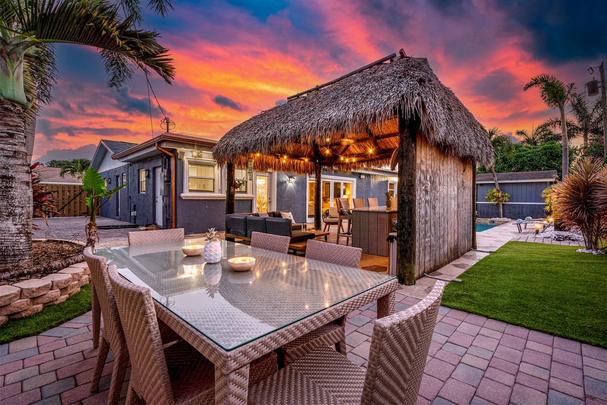 An outdoor dining area is set under a thatched-roof tiki hut, illuminated by soft string lights. A large table with ample seating is surrounded by lush greenery and landscaped stone paths, contributing to an inviting atmosphere as the sky is painted in vibrant sunset hues.