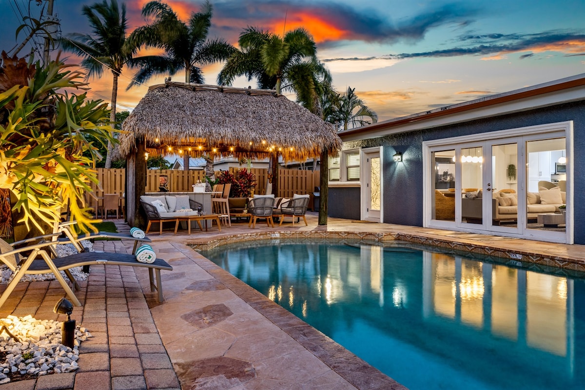 A tranquil backyard features a heated pool, surrounded by stone pavers and lush greenery. An authentic Tiki hut with warm lighting offers seating and a glimpse of the inviting indoor space through glass doors. The sunset sky complements the peaceful atmosphere.