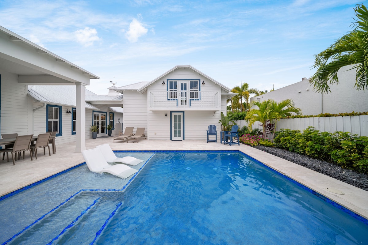 A clear view of the heated outdoor pool is presented, featuring built-in loungers on one side. The spacious patio is bordered by vibrant tropical landscaping and includes multiple seating areas. The white home is visible in the background, along with a second-floor balcony.
