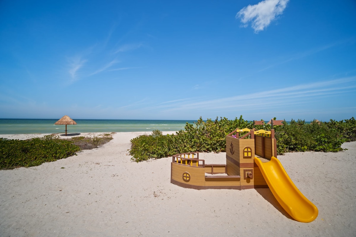 A colorful children's play structure resembling a pirate ship is situated on soft white sand, highlighting a family-friendly beach atmosphere. In the background, a thatched umbrella provides shade, while the serene turquoise waters extend to the horizon under a clear blue sky.