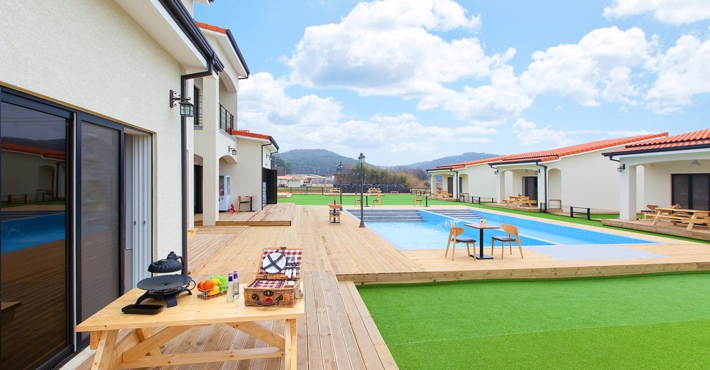 An outdoor area is showcased with a wooden deck surrounding a swimming pool. Lounge chairs are positioned by the water, and a dining table is set nearby. Green grass complements the wood, while buildings with red-tiled roofs provide a backdrop under a clear blue sky.