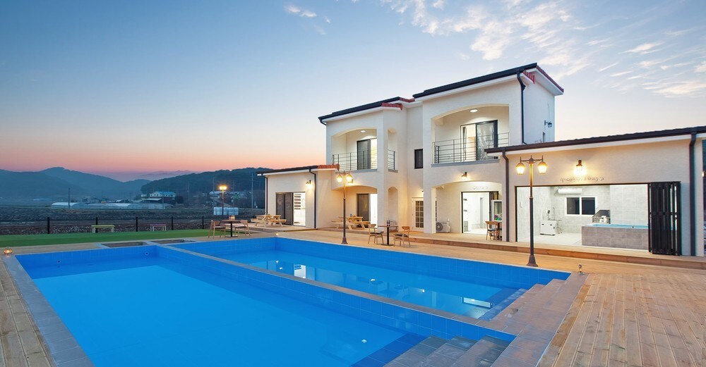 An outdoor area features two swimming pools framed by wooden decking, with lounge chairs placed nearby. The two-story building showcases large windows, allowing natural light to illuminate the interior. Dusk casts a soft glow over the scene, enhancing the tranquil atmosphere.