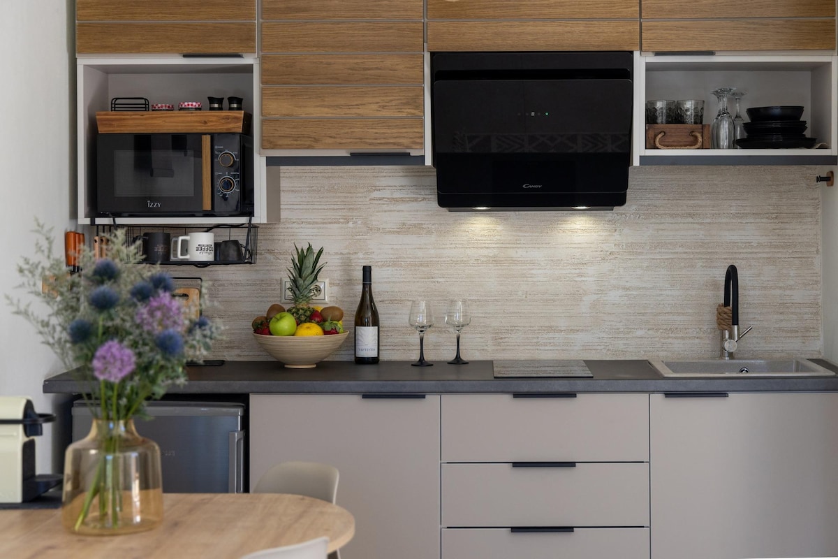 A modern kitchen is presented with wood and neutral cabinets. A bowl of fresh fruit is displayed on the counter, alongside a wine bottle and two glasses. Kitchen appliances, including a microwave and coffee maker, are neatly arranged, contributing to a functional cooking space.