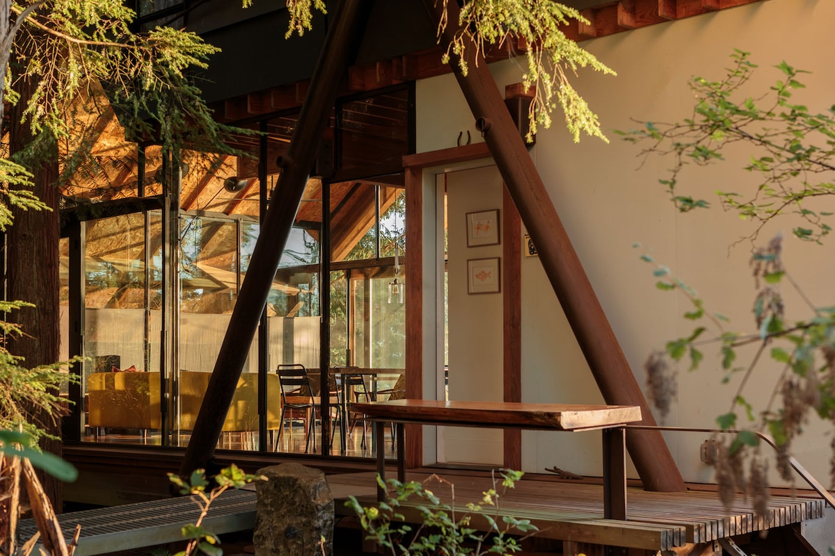An exterior view of the cabin highlights the contrasting wood and glass structure. The glass walls reflect the surrounding greenery, while the entryway displays two framed artworks. A wooden deck extends outward, providing a seamless connection to the nature beyond.