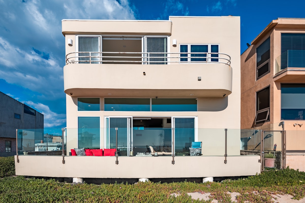 A modern beachfront home is depicted with large glass windows reflecting the sky. A glass railing encloses an outdoor seating area furnished with colorful cushions. The sandy beach and ocean are visible beyond the property, emphasizing its coastal location.