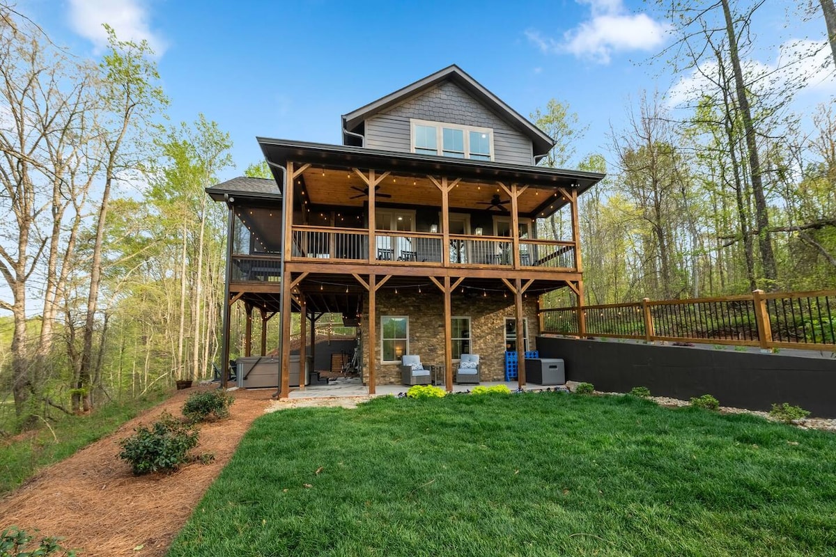 The exterior of the three-story house features a large wooden deck supported by stone pillars, surrounded by a well-kept lawn and trees. Upper and lower outdoor seating areas overlook the scenic landscape, offering ample space for relaxation and enjoyment of the natural surroundings.