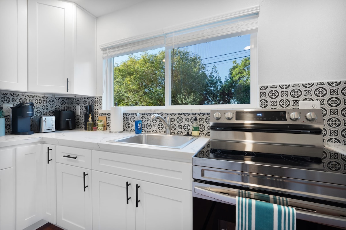 The kitchen showcases modern white cabinetry, contrasting with decorative black and white tile backsplash. Natural light fills the room through a large window, illuminating stainless steel appliances, including a stove and refrigerator. Essentials are neatly arranged on the countertops.