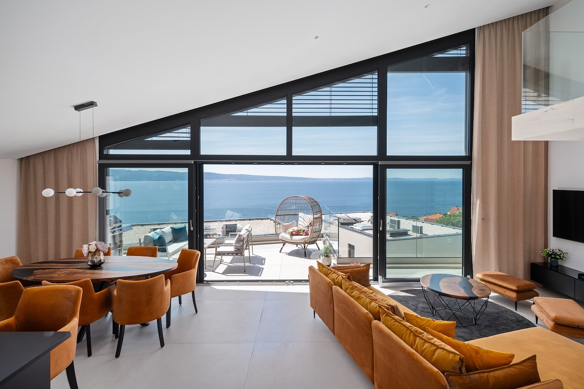 The living area features modern furnishings, including a round dining table and a cozy sofa. Large glass windows frame stunning sea views, with natural light illuminating the space. Soft curtains accentuate the contemporary design, and a stylish lounge chair adds comfort.