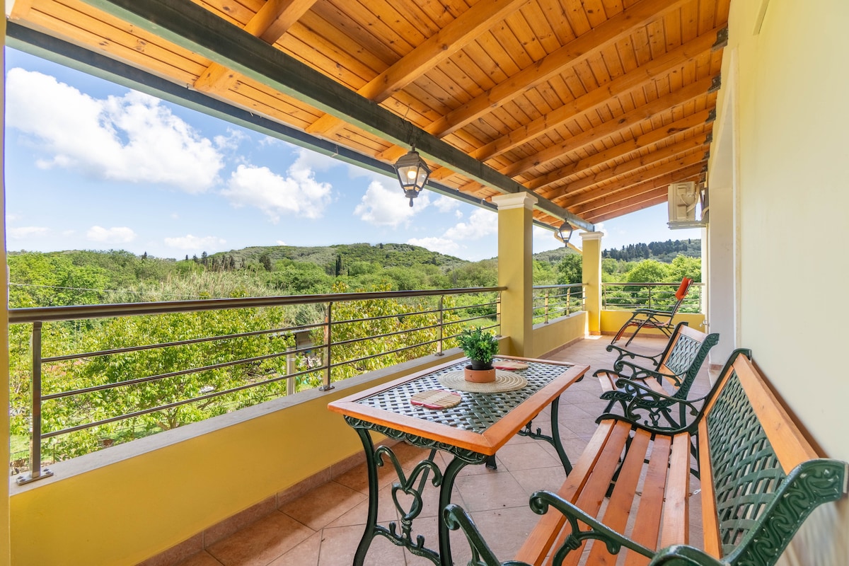 A spacious balcony features a wooden dining table surrounded by green metal chairs. Sunlight filters through the wooden beams of the roof, illuminating the verdant landscape and distant hills visible beyond the railing. An inviting atmosphere is created by the surrounding natural scenery.