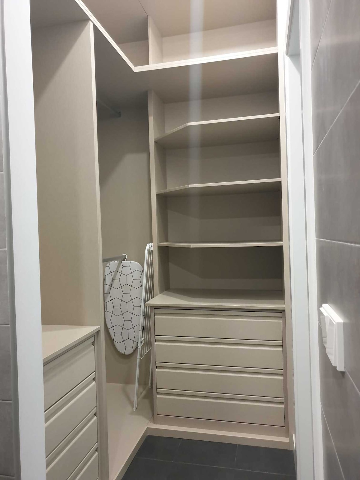 A spacious walk-in closet is presented, featuring built-in shelves and drawers for ample storage. A laundry basket is visible, alongside an ironing board. The closet's neutral-colored walls contribute to a clean and organized appearance.