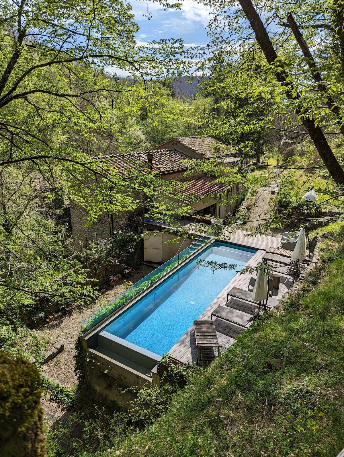 An elevated view of the hydrolysis pool is presented, surrounded by lush greenery and trees. Sun loungers and umbrellas are positioned by the poolside, inviting relaxation. The charming exterior of the Mill is visible in the background, blending harmoniously with the natural landscape.