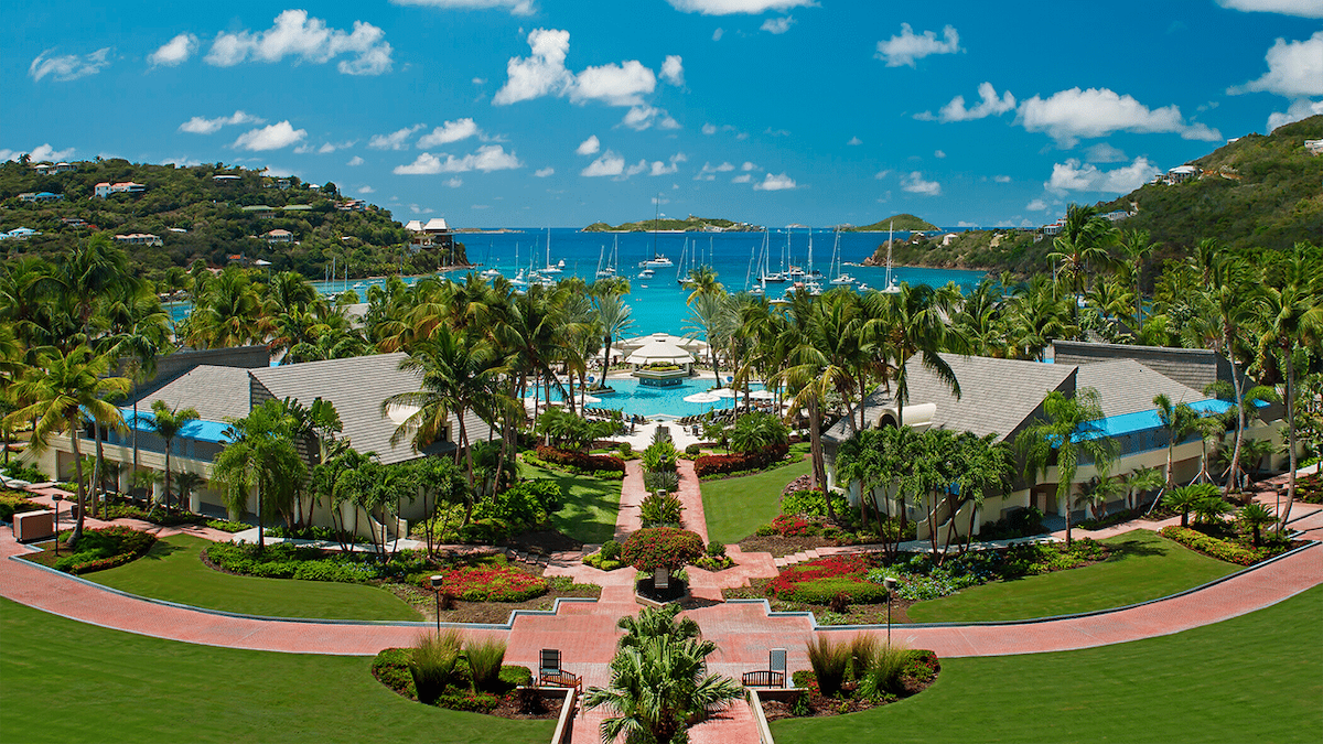 The image showcases a lush tropical landscape with manicured gardens leading to a clear blue bay. Resort buildings are visible on either side, framed by palm trees. It reveals a serene pool area surrounded by lounge chairs, with sailboats dotting the water under a bright sky.