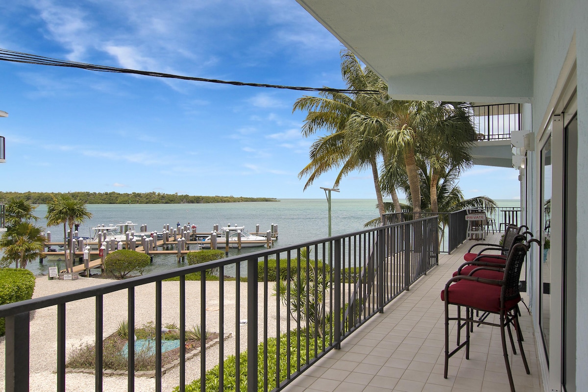 A spacious balcony extends along the waterfront, featuring several red cushioned chairs. Views of the Gulf and dock area are visible, framed by palm trees. Nearby greenery adds a touch of nature to the setting, promoting a relaxing outdoor atmosphere.