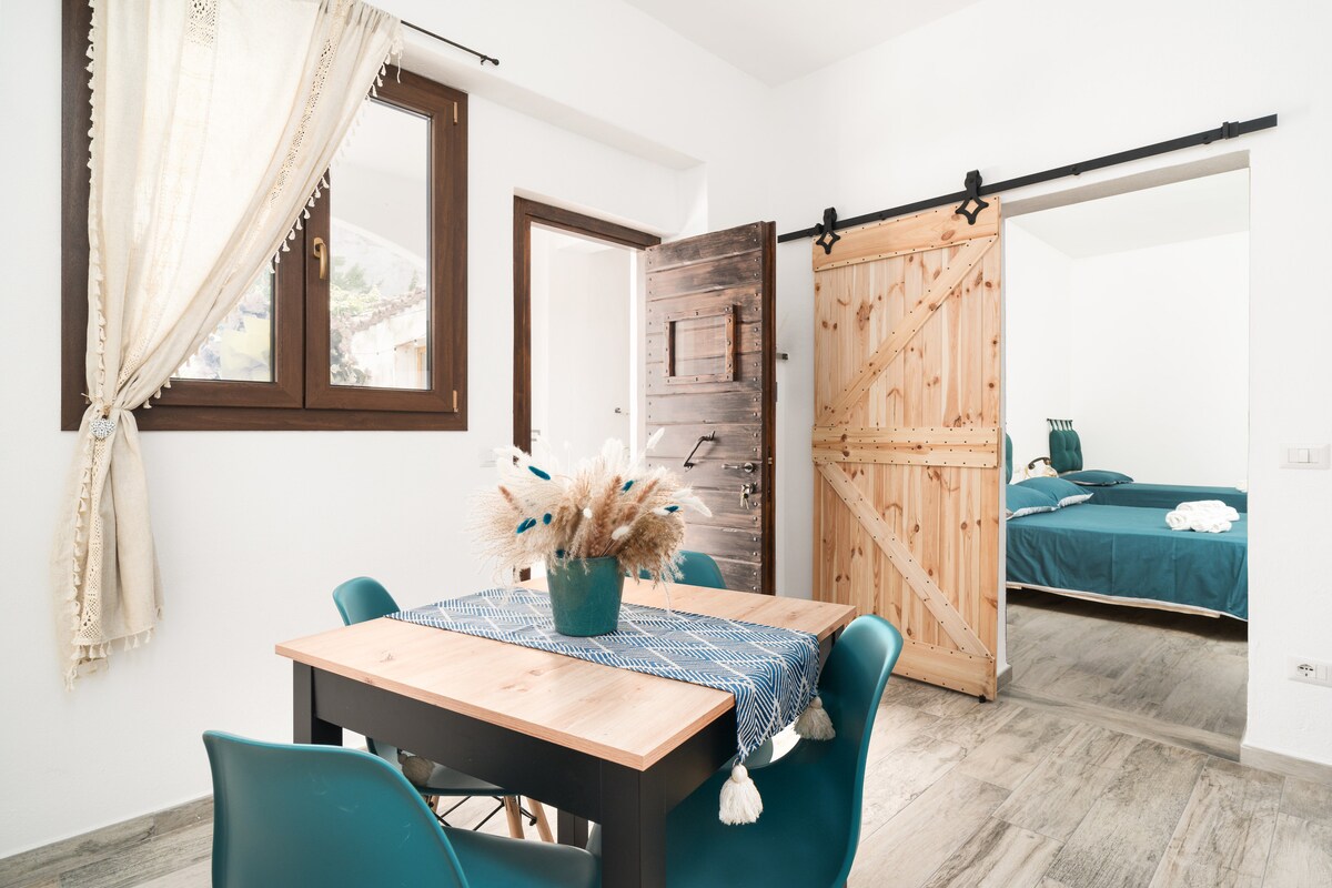 A dining area is featured with a wooden table and teal chairs. Natural light enters through a window adorned with soft curtains. The room is connected to a bedroom via a sliding wooden door, providing a glimpse of a neatly made spacious bed in calming colors.