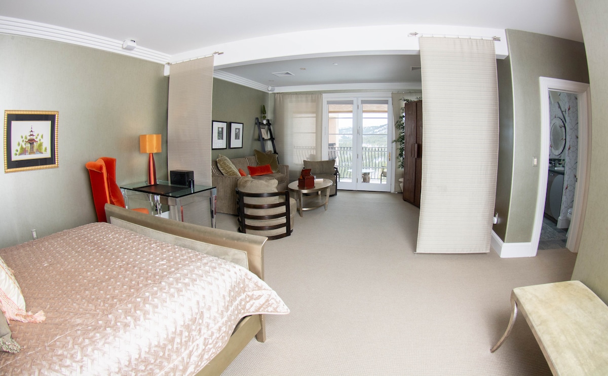 A spacious suite features a cozy sleeping area with a plush bed covered in soft linens. Comfortable seating options are arranged around a sleek glass table. Large doors open to a balcony with scenic views, while natural light filters through sheer curtains.
