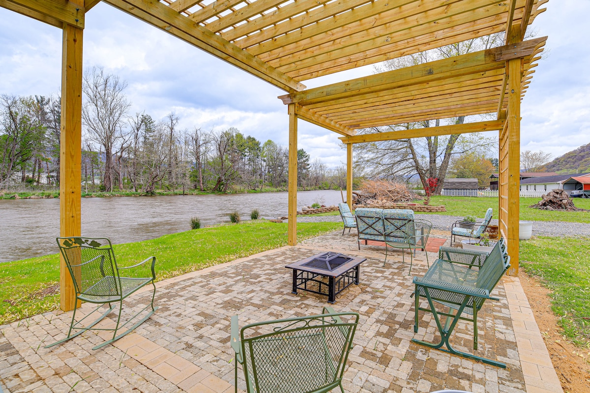A riverside pergola offers a shaded gathering space with multiple metal chairs arranged around a central fire pit. A stone patio extends beneath the structure, framing views of the Watauga River and surrounding greenery.