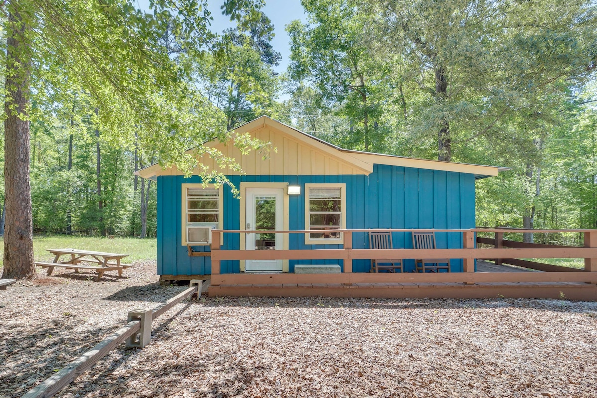 The exterior of the cabin is painted in a vibrant blue, framed by rich green trees. A covered porch features two rocking chairs, and a picnic table is situated nearby on a gravel area, creating a welcoming outdoor space.