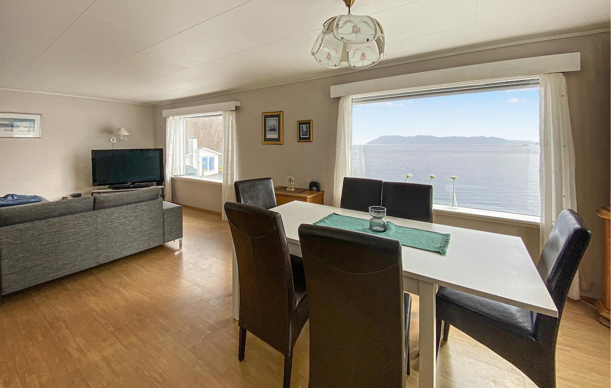A spacious living area is observed, featuring a large table with six dark chairs and a comfortable sofa. Two wide windows allow natural light to fill the room, framing views of the sea and distant hills. Simple artwork adorns the walls, enhancing the serene atmosphere.
