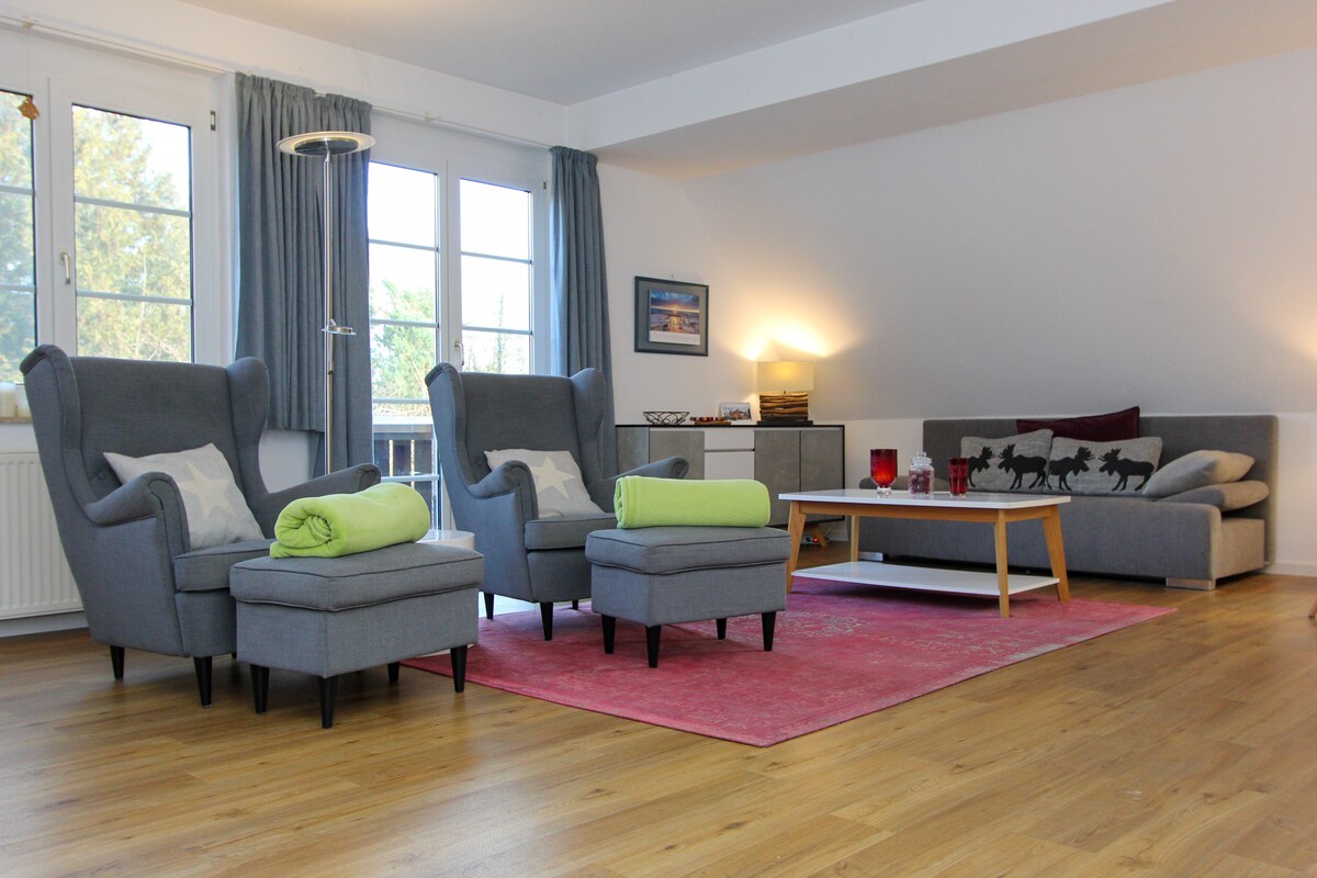 A spacious living room features two comfortable armchairs and a matching footstool, arranged around a modern coffee table. A cozy couch accommodates additional seating. Natural light fills the room through large windows, complemented by light-colored walls and a vibrant area rug.