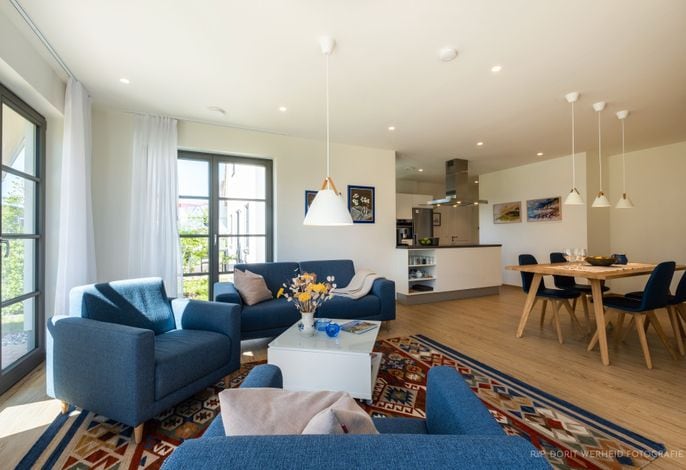 A bright living area is furnished with blue sofas and a central coffee table, surrounded by large windows that allow natural light. A dining table is set for four, and art pieces adorn the walls, enhancing the modern interior design.