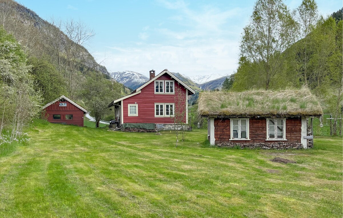 A scenic view of a red holiday home is presented, accompanied by two additional structures. Lush green grass covers the area, with trees visible in the background. The mountains in the distance are partially capped with snow, adding to the serene landscape.