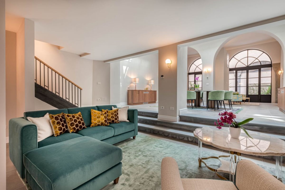 A spacious living area is adorned with modern furnishings, featuring a large teal sectional sofa embellished with patterned cushions. Arched doorways lead to an elegantly designed dining area, illuminated by soft lighting, with green upholstered chairs and a wooden sideboard in the background.