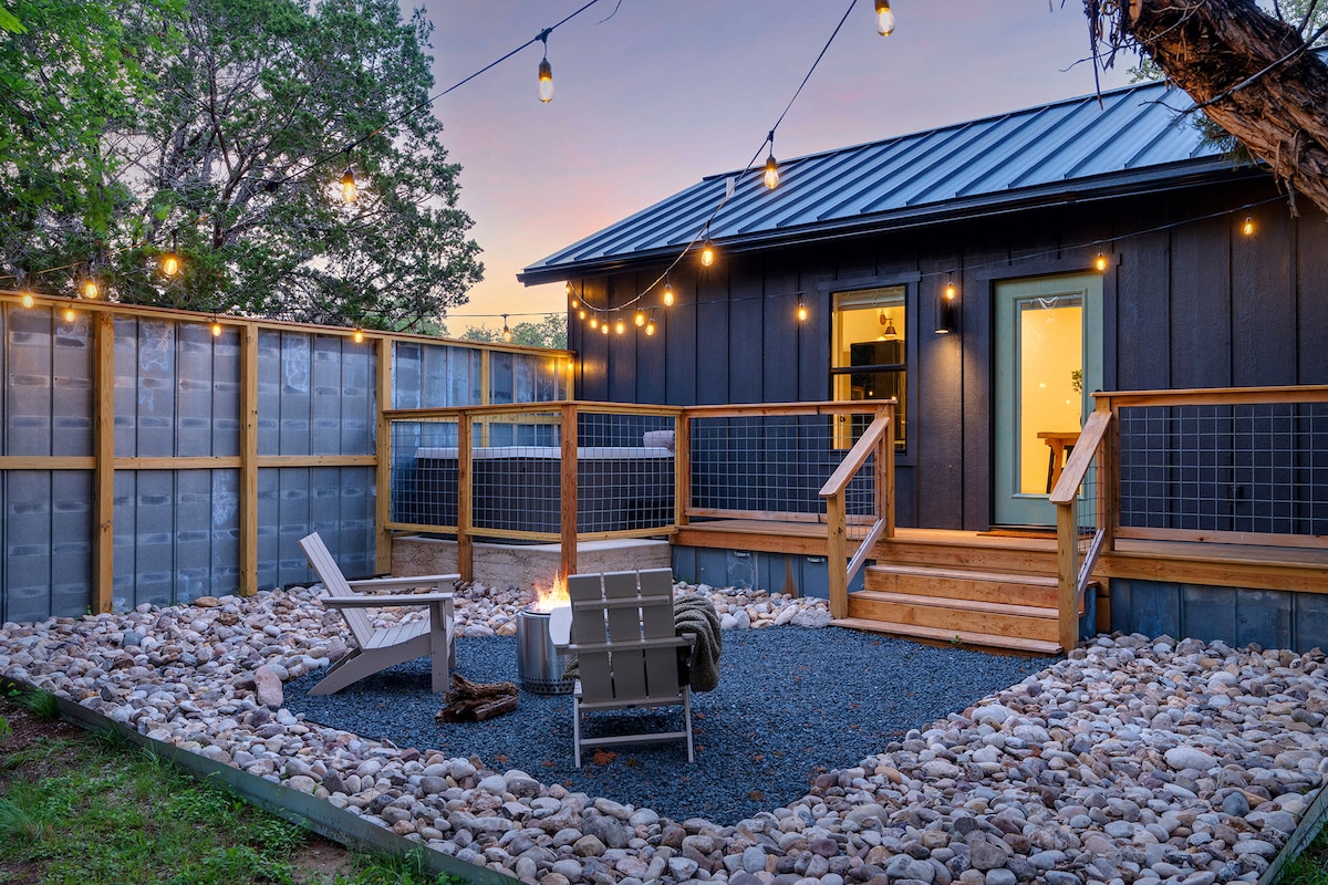 An outdoor area is illuminated by string lights, featuring a set of comfortable chairs surrounding a stone fire pit. The wooden deck leads to the entrance of the cottage, with natural stone landscaping enhancing the serene ambiance.