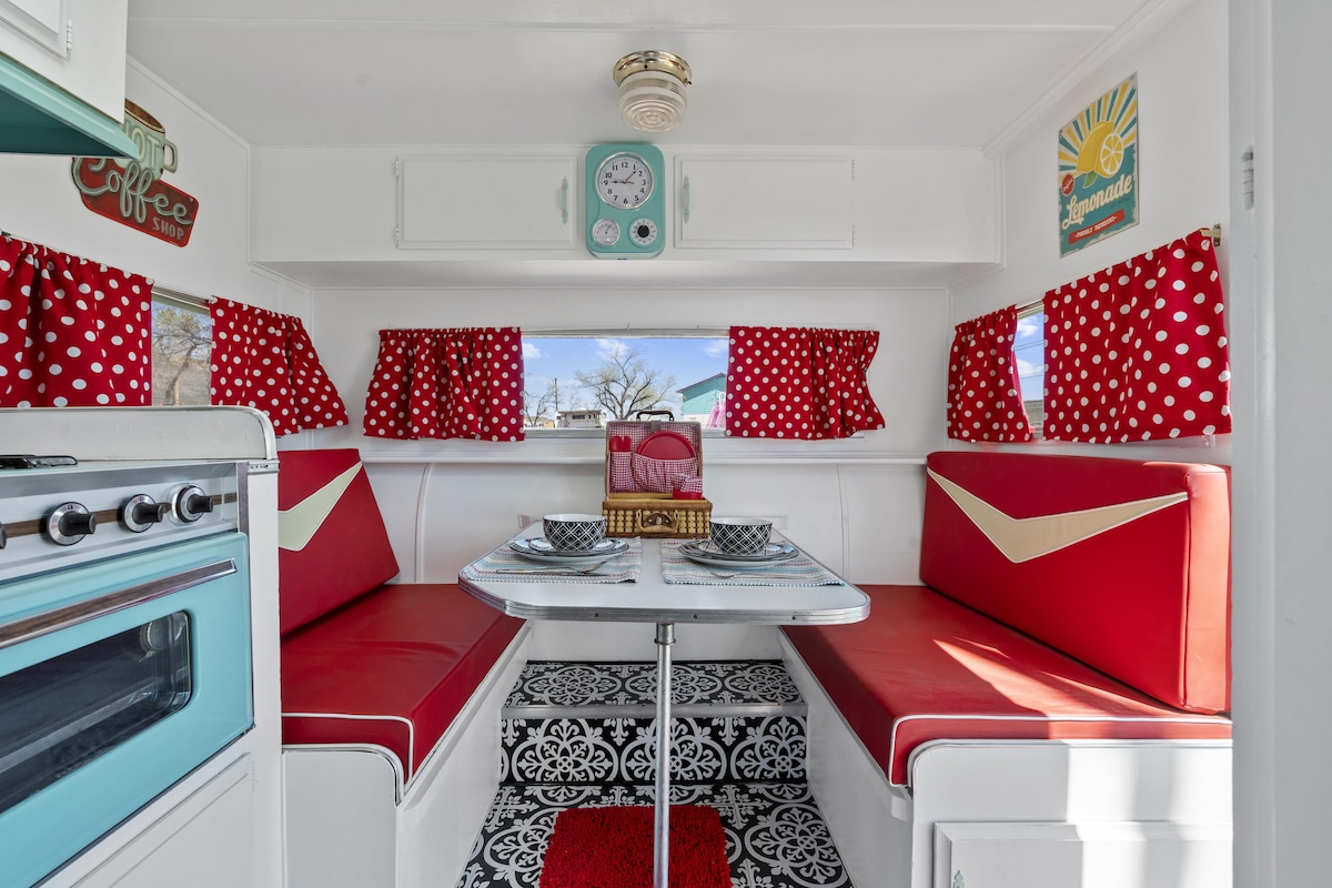 A cozy dinette area features retro red and white polka dot curtains and seating, with a round table set for two. A vintage stovetop and refrigerator can be seen in the foreground, creating a charming 1960s dining atmosphere.